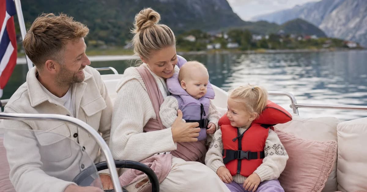 Familie i fritidsbåt på fjorden med småbarn iført rød redningsvest tilpasset barnestørrelse