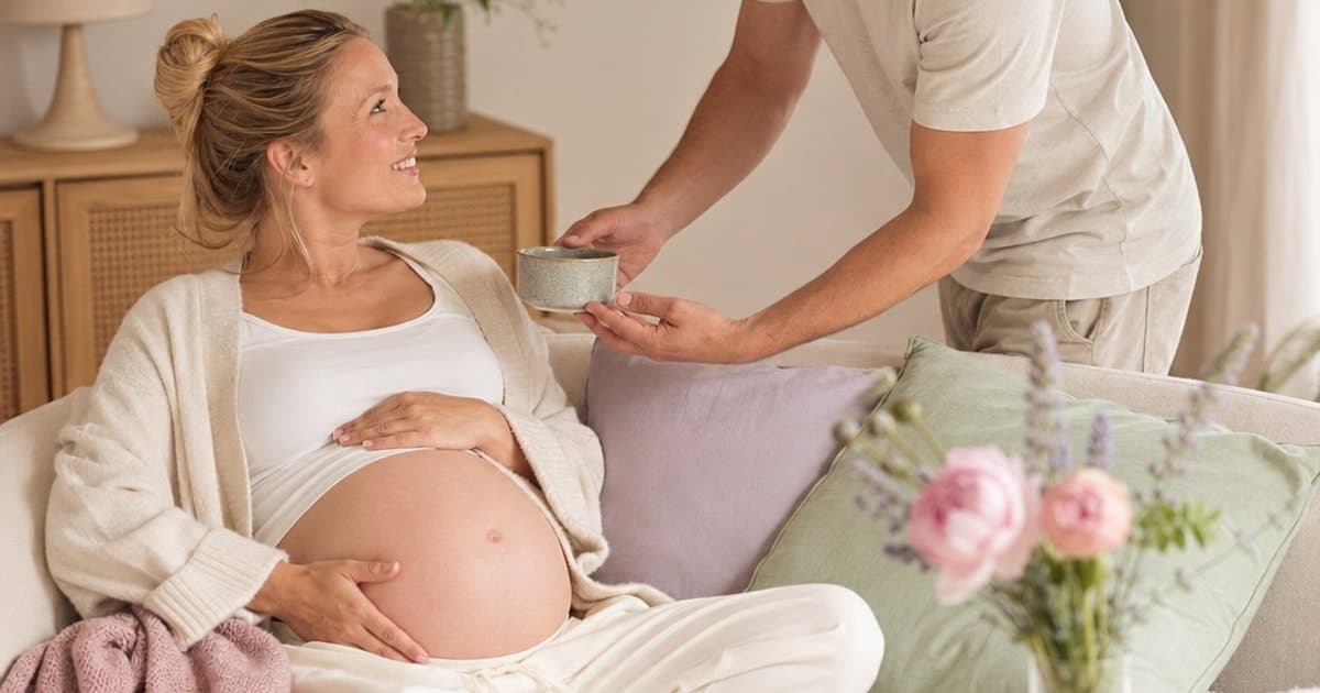 Gravid kvinne med stor mage hviler hjemme mens partneren bringer henne en kopp te