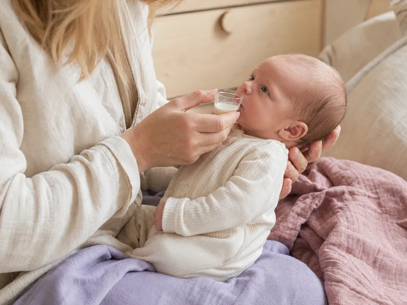 Nærbilde av et lite medisinbeger med morsmelk som holdes mot en babys underleppe