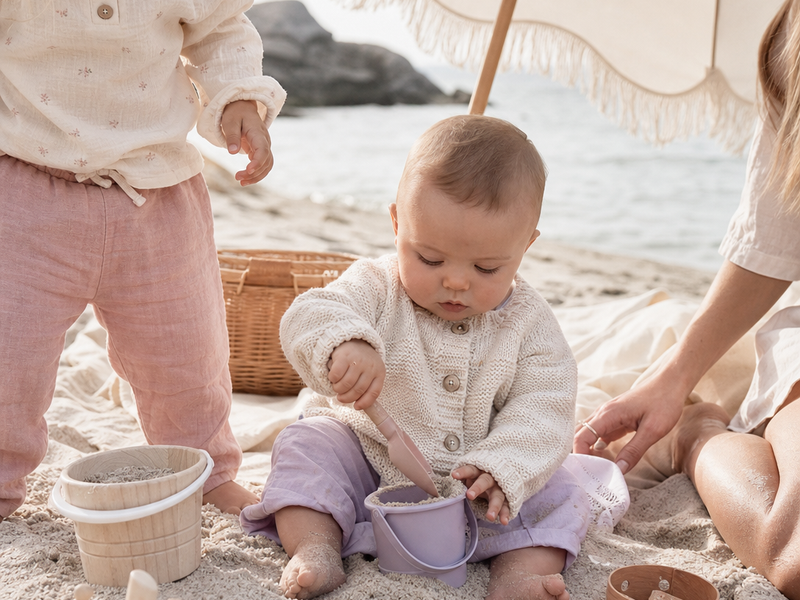Baby leker trygt i skyggen av et strandtelt med bøtte og spade på sanden